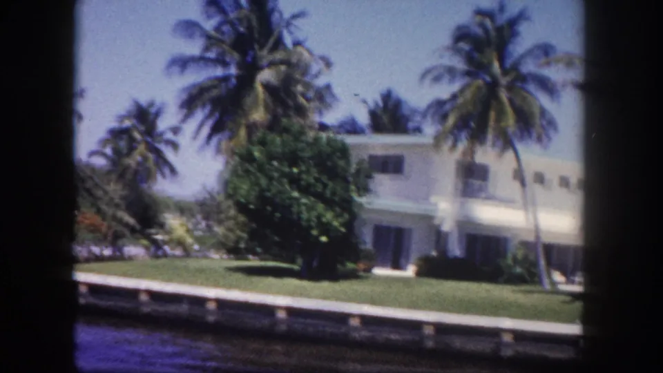 8mm film frame circa 1960 of a white flat-roofed Florida waterfront residence amid palm trees and tropical vegetation, shot from a boat on a Tampa-area canal.