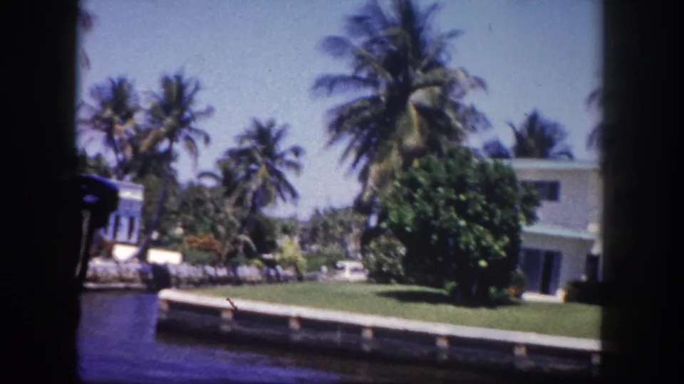 Canal view of a flat-roofed white residence with lush palm trees on a sunny day, Tampa, Florida, circa 1960, 8mm home movie.