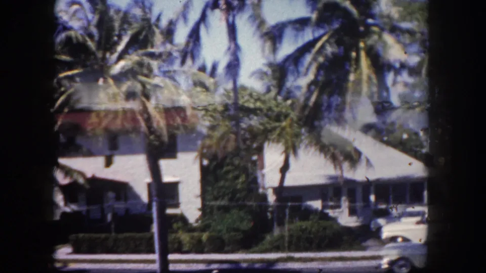 White residential building with red-tiled roof framed by palm trees, parked classic sedan at lower right; 8mm home movie, Tampa, Florida, circa 1960.