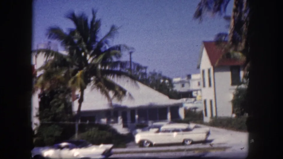 Light-colored classic American sedan on a sunny street flanked by white buildings and coconut palms; 8mm home movie, Tampa, Florida, circa 1960.