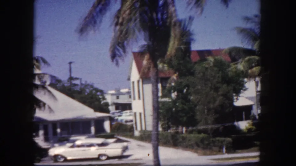 White classic American sedan near white gabled buildings with red roofs, framed by palm trees; 8mm home movie, Tampa, Florida, circa 1960.