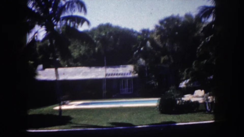 8mm home movie frame, c.1960, Tampa, Florida: backyard swimming pool edged by green lawn, tall palms, and a ranch-style house with tiled roof.
