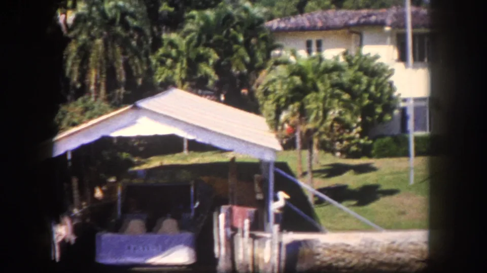 8mm home movie, c.1960, Tampa, Florida: covered boat dock or boathouse with white roof, tropical vegetation, stucco house in background, small figure discernible near dock.