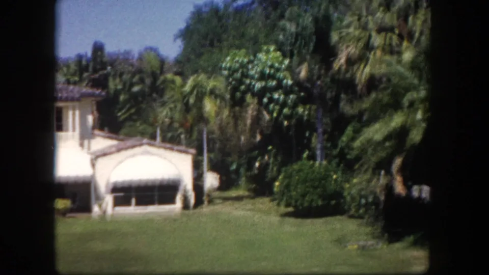 Right end of Mediterranean-style house with palm trees and tropical foliage foregrounded, 8mm home movie, Tampa Florida area, c.1960.