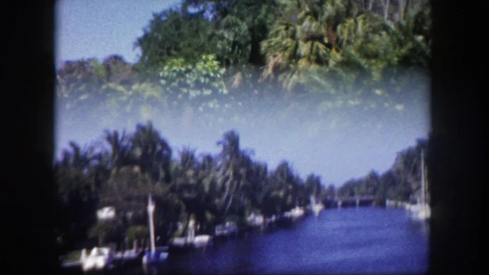 8mm film frame of a palm-lined canal in Tampa, Florida, circa 1960; sailboat mast and mooring structure on left bank under a clear blue sky.