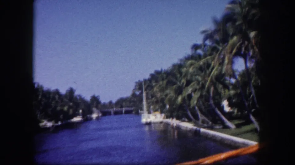 8mm film frame of a Tampa, Florida canal circa 1960; a low bridge appears in the middle distance with leaning palms and moored sailboats on the right bank.