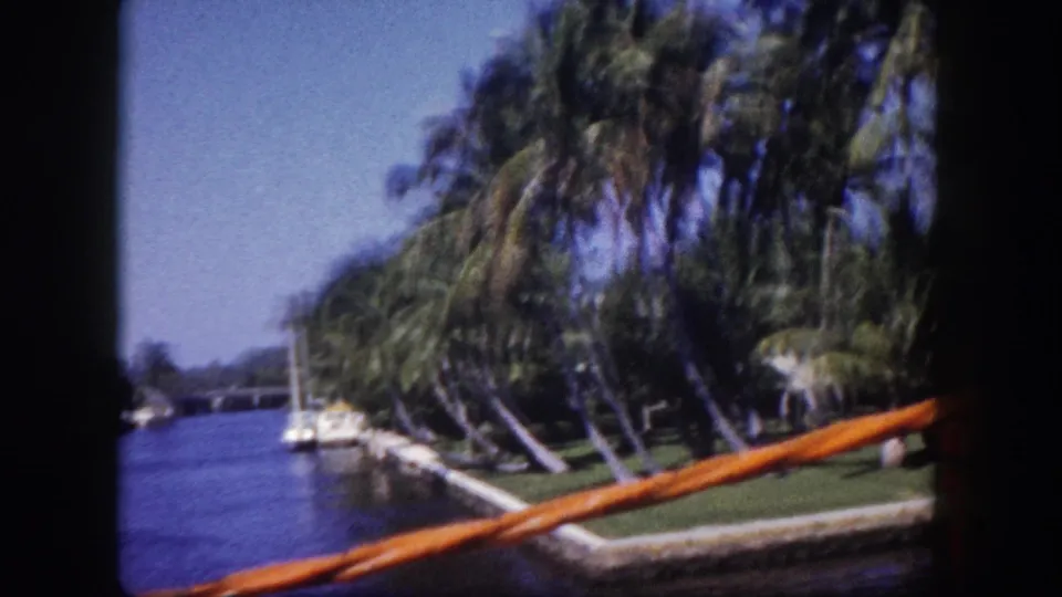 8mm film frame shot from a moving boat on a Tampa, Florida canal circa 1960; orange boat gunwale in lower right, leaning palms, moored sailboats, and a bridge visible ahead.