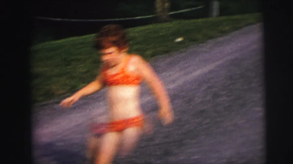 8mm home-movie frame of a young girl in a red bikini running on a gravel path, circa 1976, with characteristic motion blur from handheld 8mm capture.