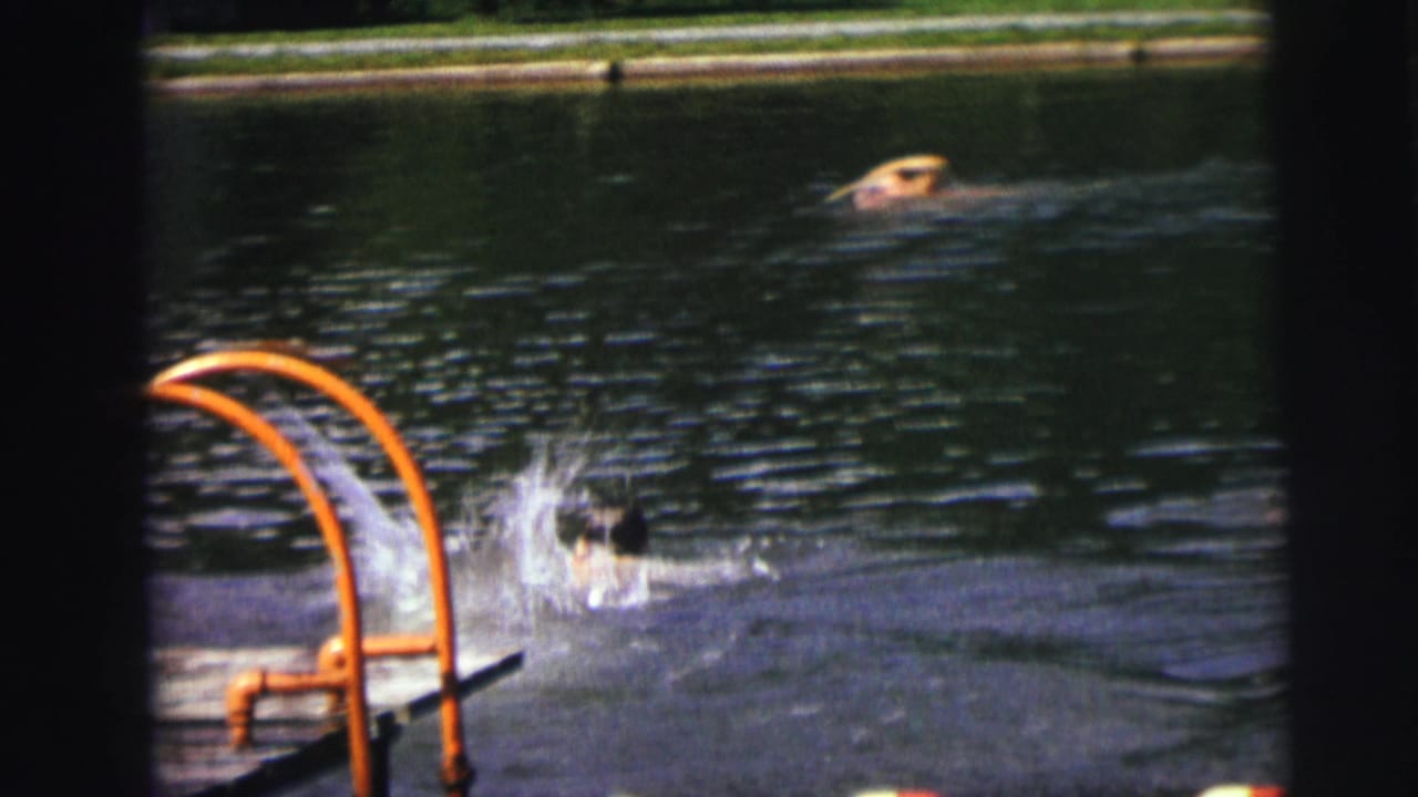 Swimmer doing an energetic freestyle stroke in a blue-grey outdoor pool, orange tubular metal pool ladder prominent at center-left.