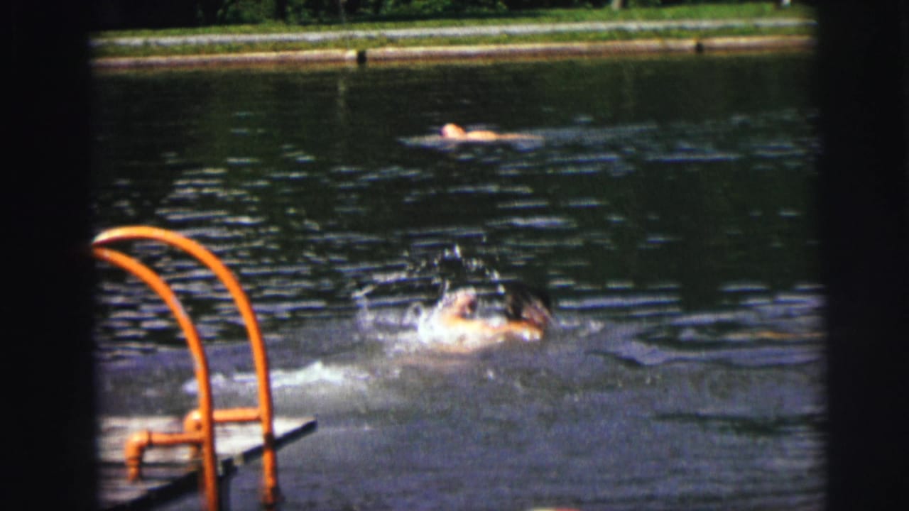 Swimmer splashing near orange pool ladder with a second swimmer visible floating in the upper background.