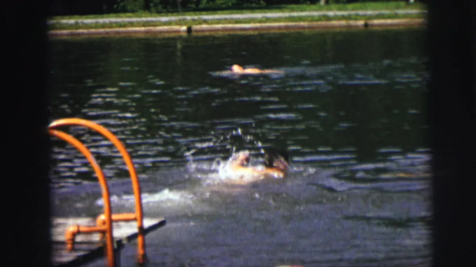 Two swimmers in a backyard outdoor pool — one actively stroking near the orange ladder, a second figure floating in background, c.1976 8mm.