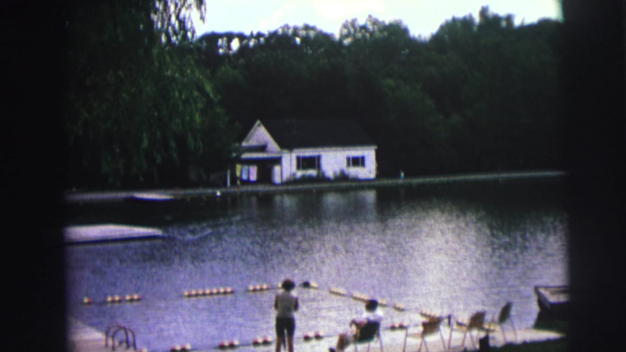 8mm home movie frame from 1976 showing a calm lake, white boathouse on the far shore, orange buoy float line, and figures on the grassy bank in Lynbrook, New York.