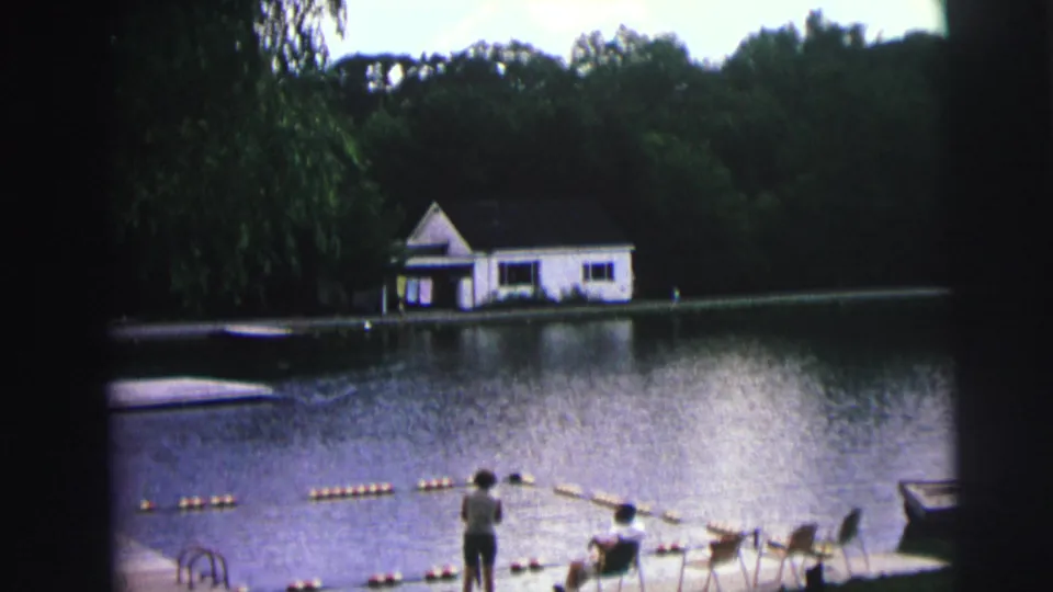 8mm home movie frame from 1976 showing a calm lake, white boathouse on the far shore, orange buoy float line, and figures on the grassy bank in Lynbrook, New York.
