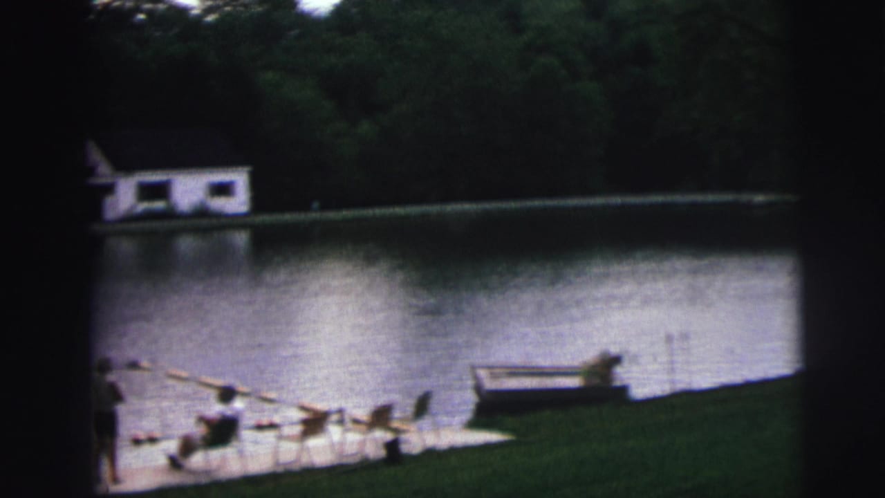 8mm home movie frame circa 1976 of a lake recreation area in Lynbrook, New York, showing lawn-chair figures, buoy swim lane, and white boathouse across the water.