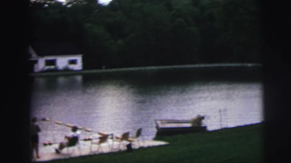 8mm home movie frame circa 1976 of a lake recreation area in Lynbrook, New York, showing lawn-chair figures, buoy swim lane, and white boathouse across the water.