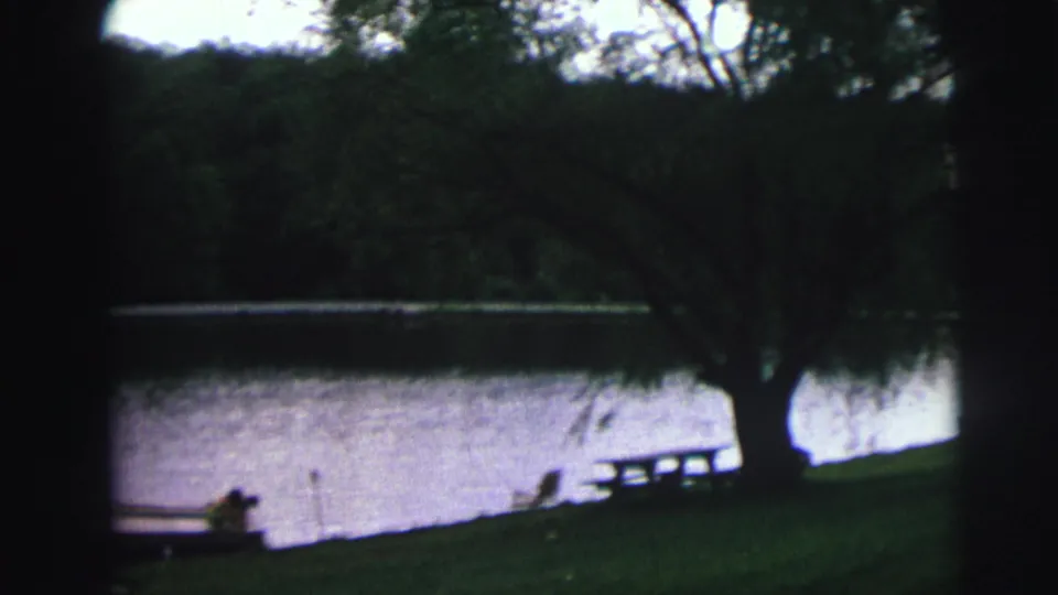 8mm home movie frame showing a shaded lakeside park with a weeping willow, picnic table, and calm lake surface, likely Lynbrook, New York, circa 1976.