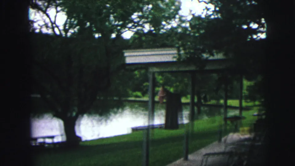 8mm home movie frame showing a tree-lined lake park with a covered structure at right, possibly Lynbrook, New York, circa 1976.
