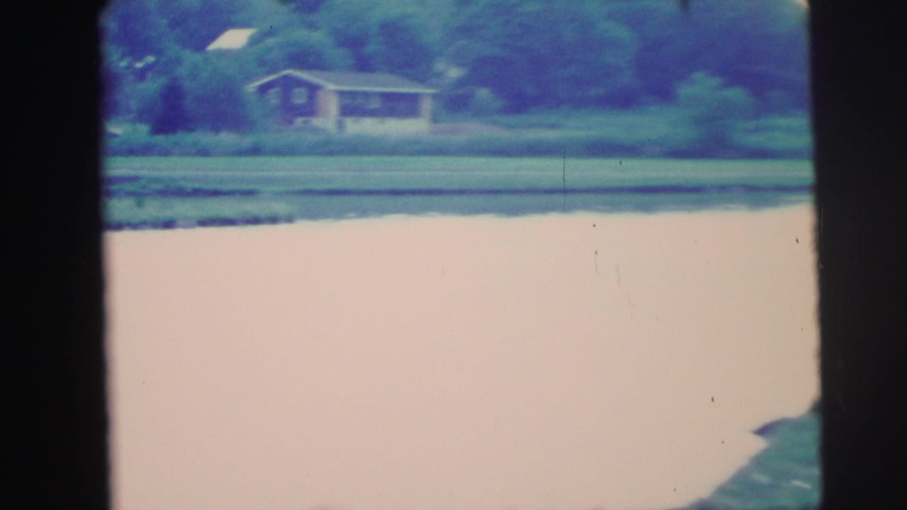 Open lake with warm pink-toned water and a house on the right shore, 8mm home movie circa 1976, Lynbrook area