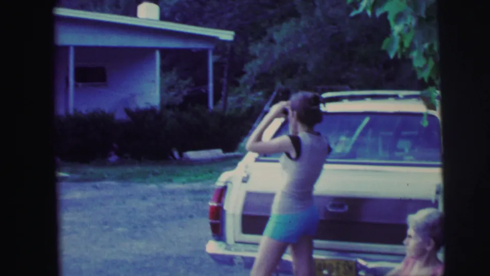 Woman adjusting hair beside a mid-1970s American station wagon, ranch house in background, 8mm home movie circa 1976