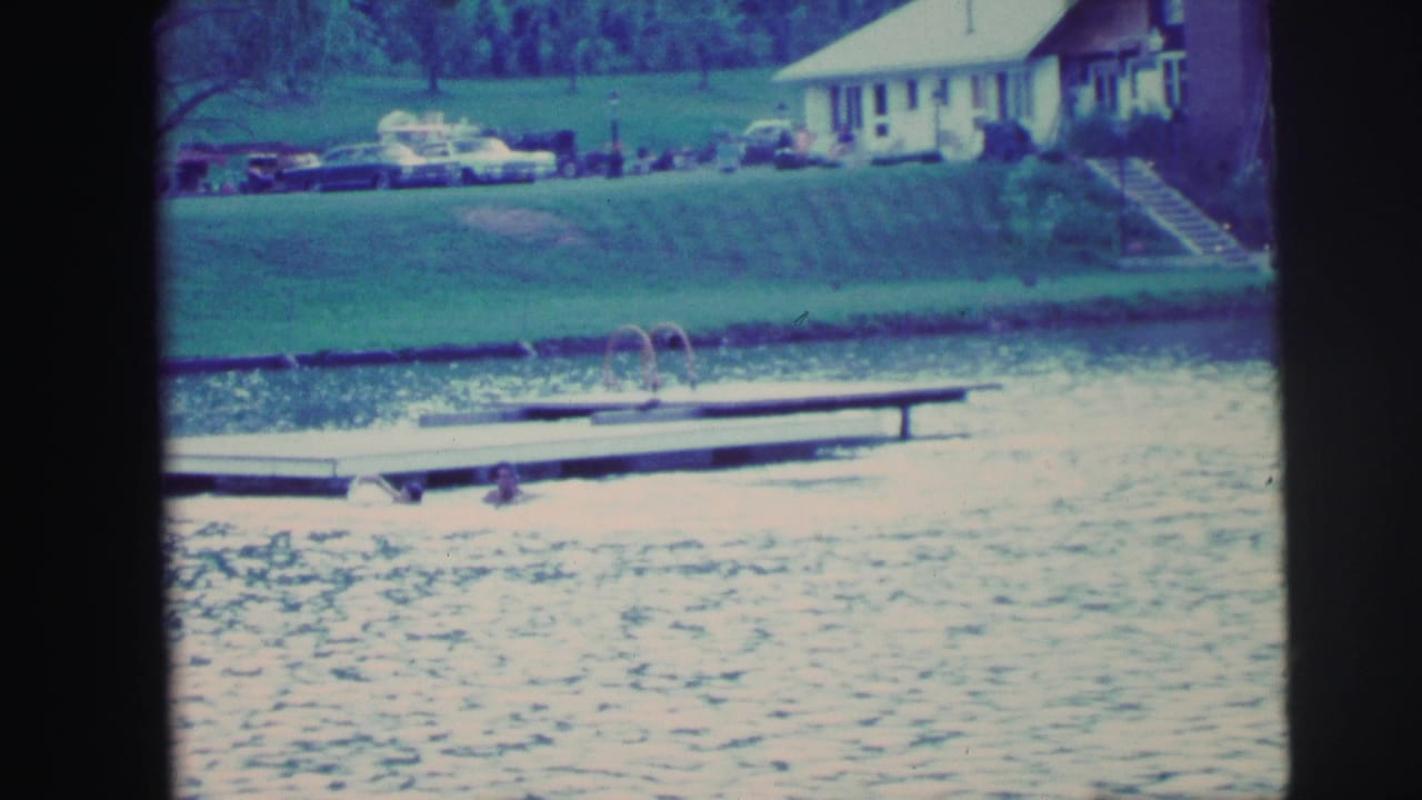 Lake view with dock visible mid-frame, multiple parked cars and a white building along the far shoreline, blue-cyan film color cast.