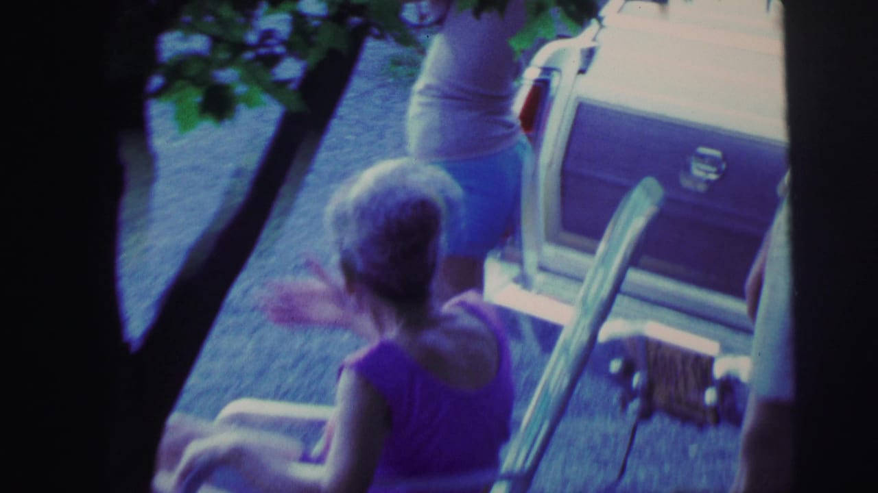 8mm home movie frame, Lynbrook, New York, 1976: gray-haired woman in purple top in lawn chair, younger woman in white tank top near station wagon, framed through tree branches