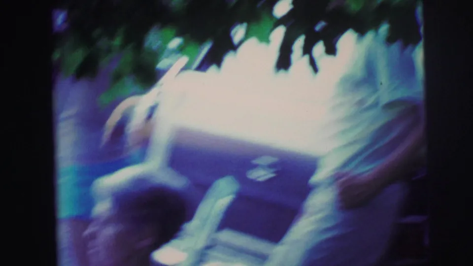 Motion-blurred 8mm film frame circa 1976: camera pan captures two women and a folding lawn chair, leafy foliage at top edge