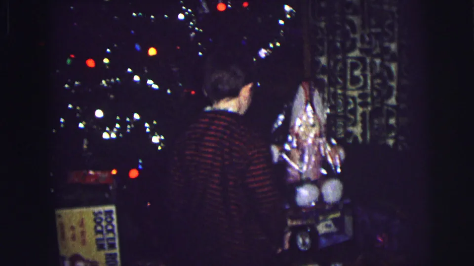 Child in plaid or striped pajama top, viewed from behind, stands at a lit Christmas tree with multicolored lights and wrapped packages on the floor; Lynbrook, New York, 1976.