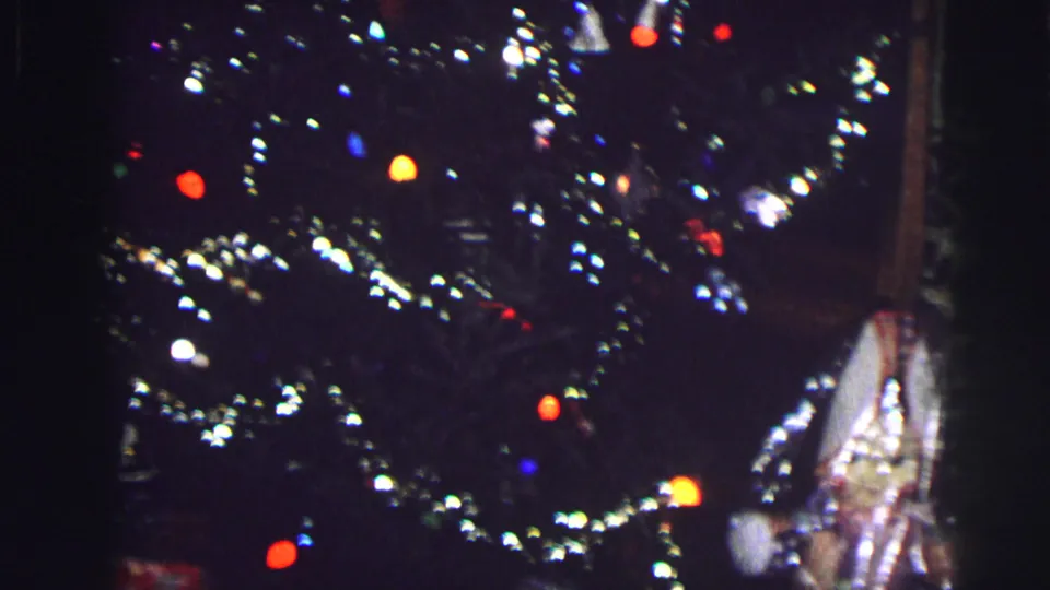 1976 home movie — Christmas tree upper section with colored incandescent lights, silver tinsel garland, and a wrapped present, wooden door frame at right edge.
