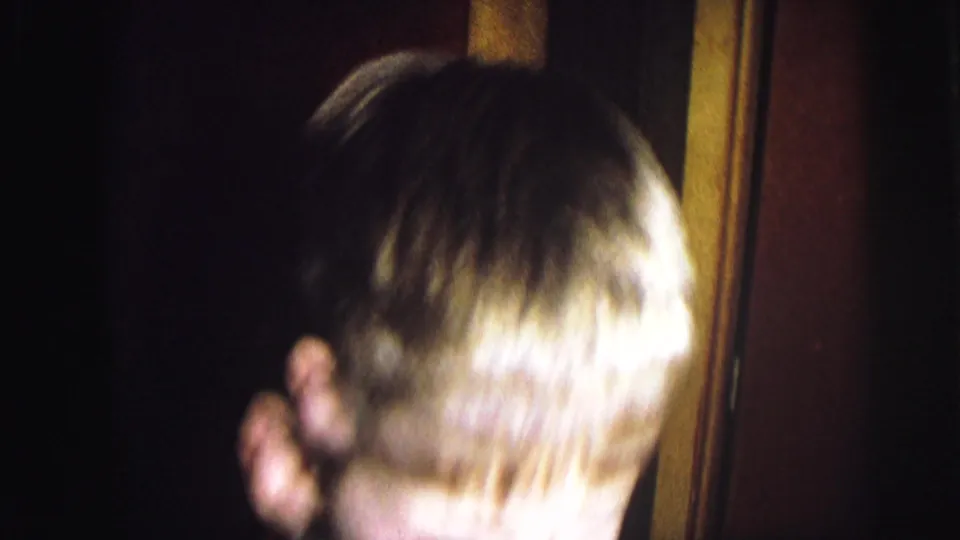 1976 home movie — young child seen from behind near a wooden door frame, apparently facing the Christmas tree area; face not fully visible.