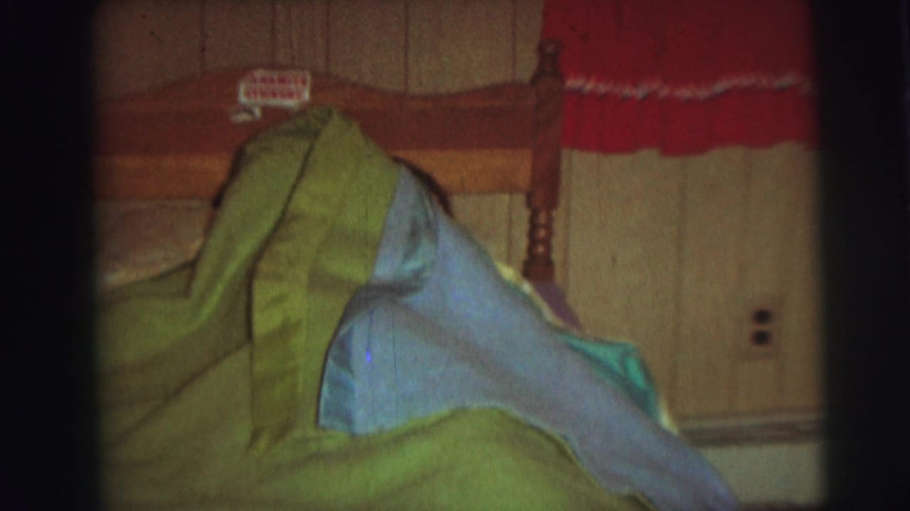 A 1976 Lynbrook, New York home-movie frame shows green and blue bedding piled on a wooden bed in a wood-paneled bedroom, with a red curtain above the headboard.