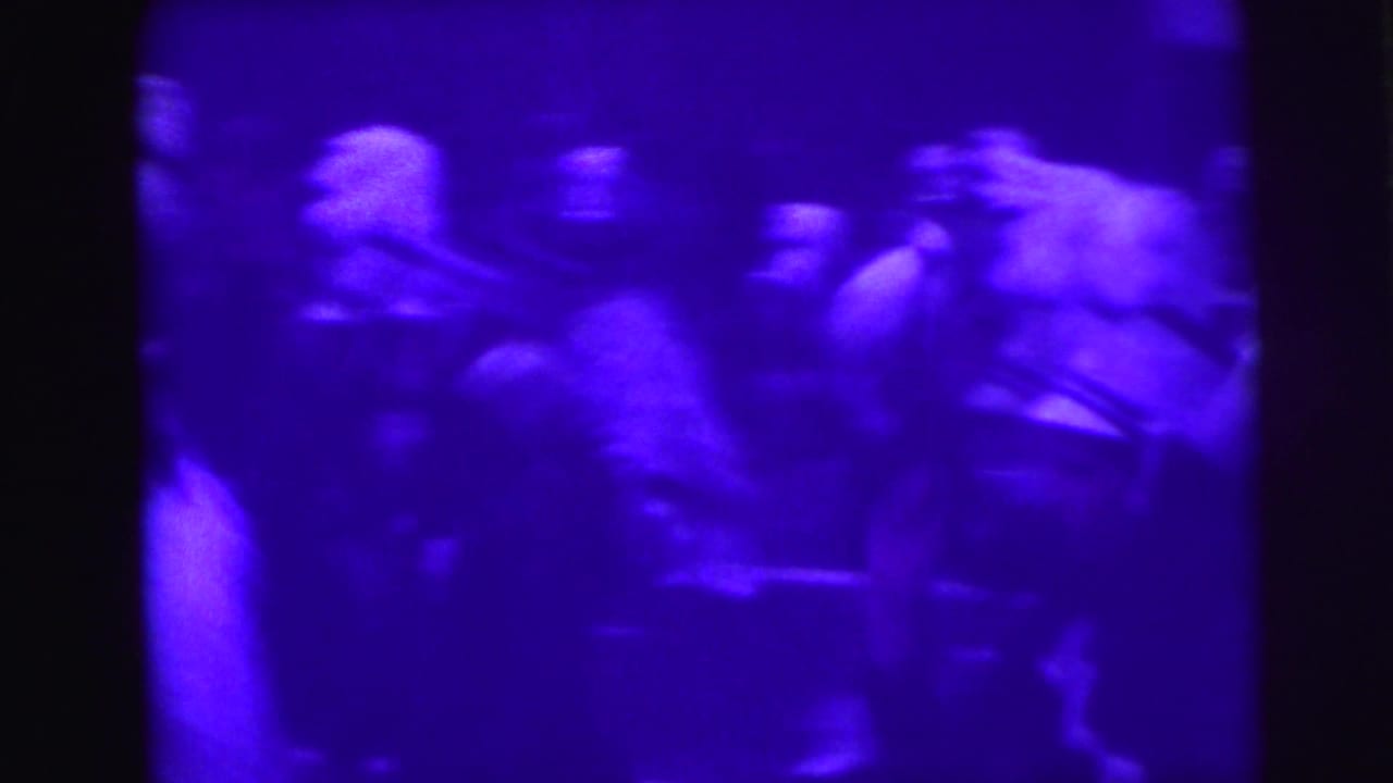 Drill team marchers in dark uniforms and white-plumed hats at close range during a 1976 Lynbrook, New York parade; heavy motion blur and purple dye-degradation tint throughout.