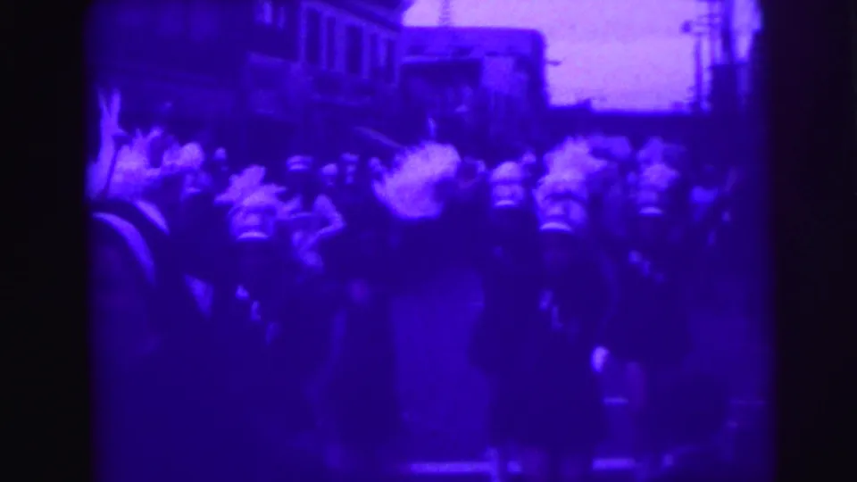 Street-level view of a 1976 Lynbrook, New York parade showing drill team marchers in dark uniforms with white plumes among spectators, with low-rise commercial buildings in the background.