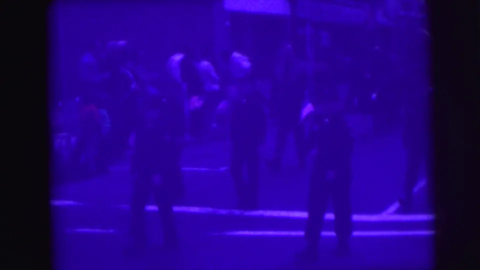 Parade participants in dark uniforms near the close of a 1976 Lynbrook, New York drill team procession; deep purple-violet cast and shadowing limit fine detail.