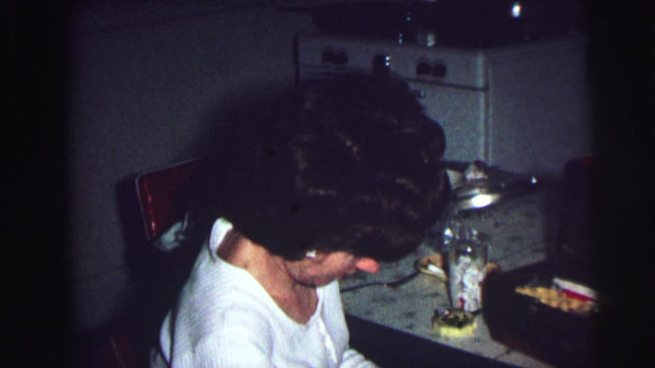 Woman with dark bouffant hair in white top leaning over a kitchen counter with glass jars and a food-filled pan.