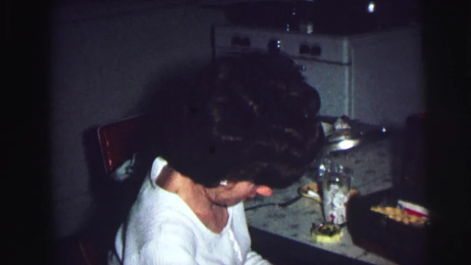 Woman with dark bouffant hair in white top leaning over a dim kitchen counter, 8mm home movie, Lynbrook NY, 1976.