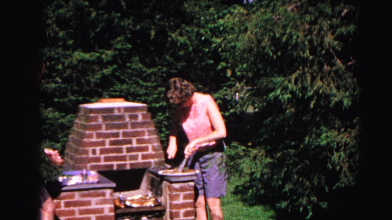 In 1963 Hagerstown home-movie footage, a woman in a pink sleeveless top and blue shorts tends food at a brick backyard grill surrounded by trees.