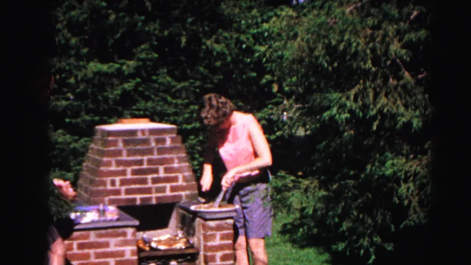 In 1963 Hagerstown home-movie footage, a woman in a pink sleeveless top and blue shorts tends food at a brick backyard grill surrounded by trees.