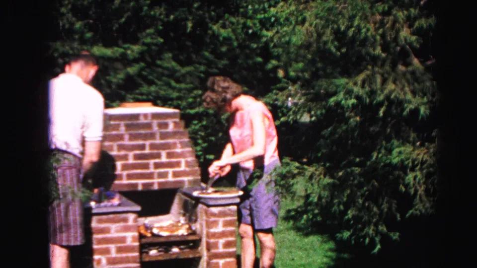 A man in a white shirt and plaid shorts joins the woman at the brick outdoor grill in the Hagerstown backyard home-movie scene.