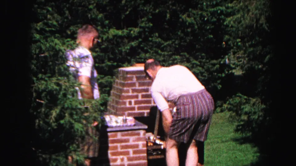 Man tends a built-in brick backyard grill while a shorter companion watches; Hagerstown, Maryland, c.1963 8mm home movie.