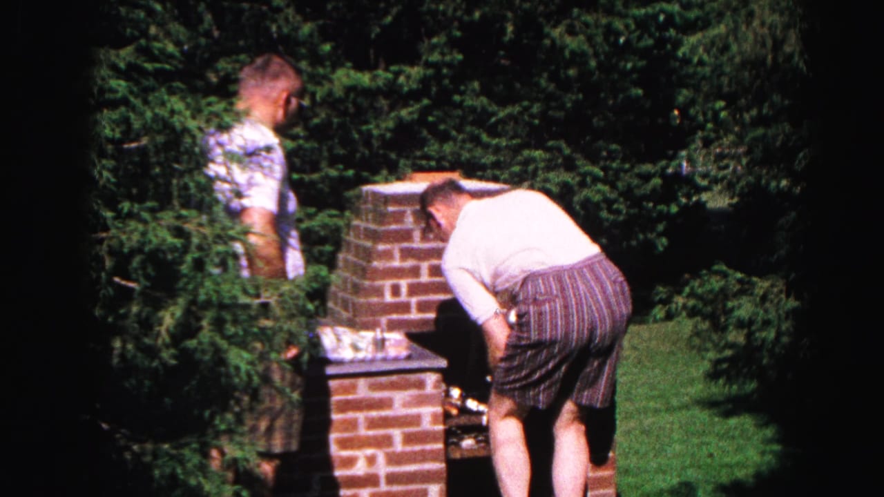 Man in white shirt leans over brick grill; second smaller figure partially visible at left; dense green foliage fills the background.