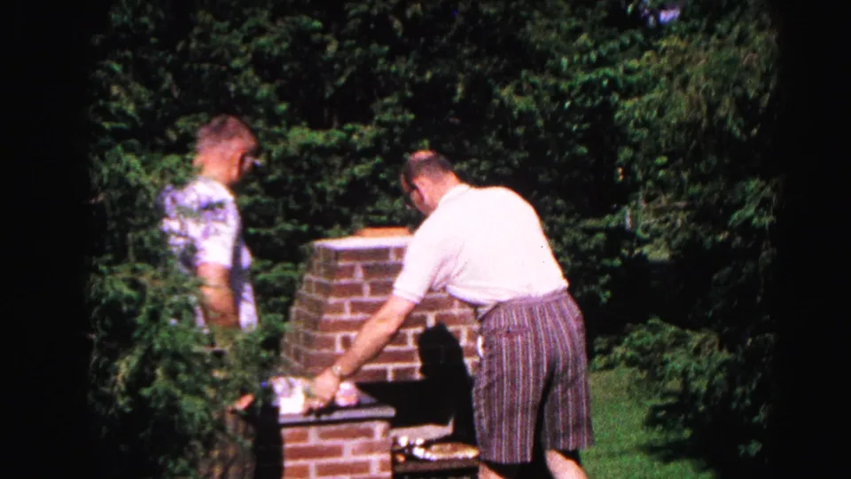 Adult man adjusts the lower firebox area of a brick barbecue while companion watches; sunlit backyard with dense evergreens, c.1963 8mm home movie.
