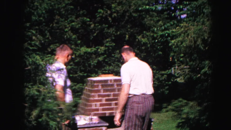 Adult man in three-quarter profile tends a brick backyard grill with a companion nearby; c.1963 8mm home movie, Hagerstown, Maryland.