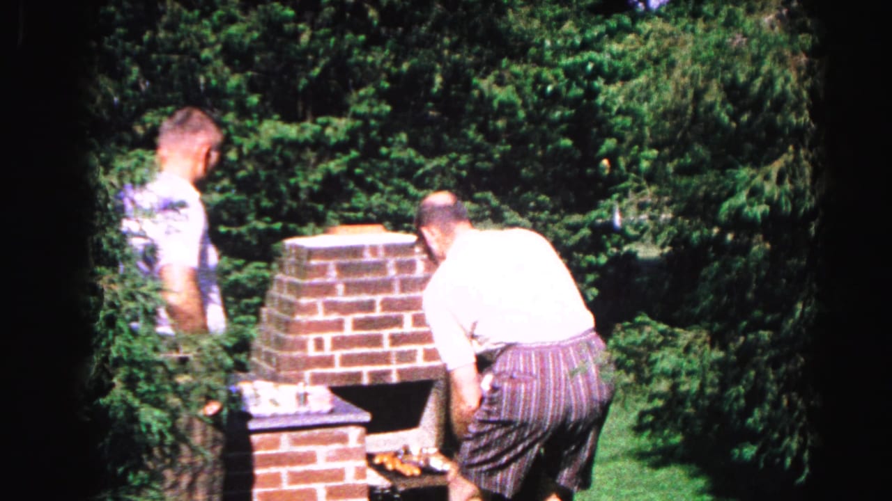 Two men in casual 1960s attire tending a tall brick backyard grill, dense deciduous trees behind, from a 1963 Hagerstown Maryland home movie.
