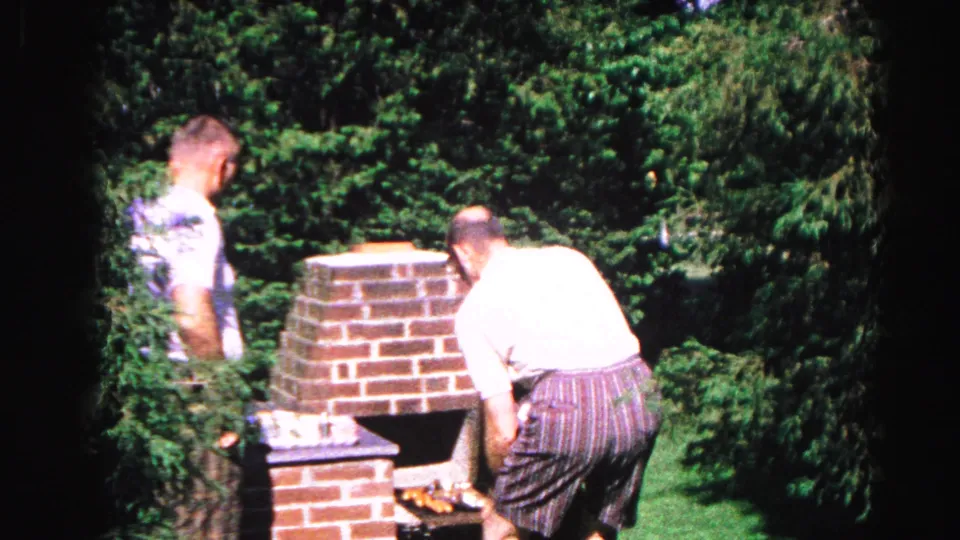 Two men in casual 1960s attire tending a tall brick backyard grill, dense deciduous trees behind, from a 1963 Hagerstown Maryland home movie.