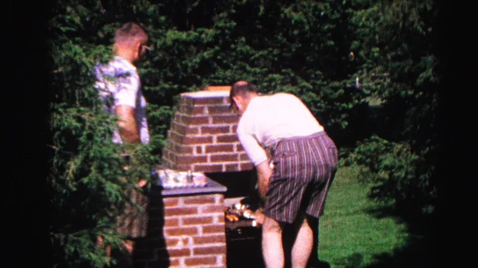 Man in striped shorts tending food on a brick backyard grill, second man visible at left, dense trees behind, from a 1963 Hagerstown Maryland home movie.