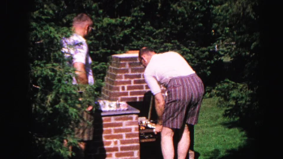 Man in striped Bermuda shorts leaning over a tall brick backyard grill, dense deciduous trees filling the background, 1963 Hagerstown Maryland home movie.