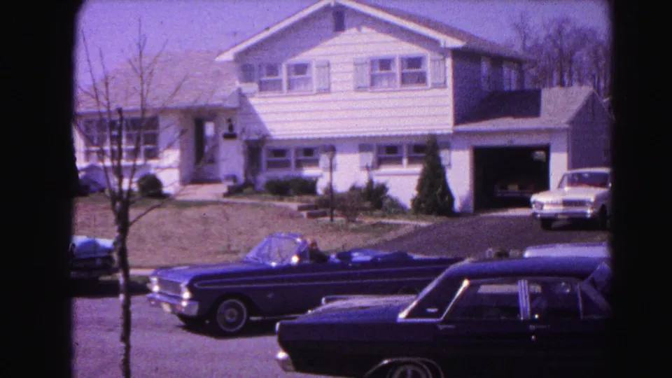 Dark blue early-1960s convertible departing a white-sided two-story suburban home with open attached garage; dark station wagon parked nearby; Hagerstown, Maryland, 1963 8mm film.