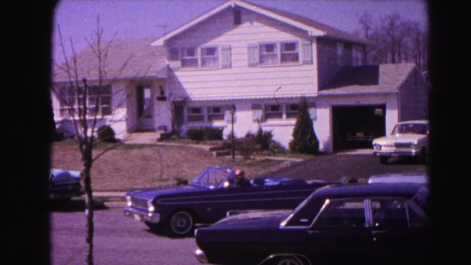 Early-1960s dark blue open-top convertible moving past a white-sided suburban home with open garage; dark station wagon and lighter sedan parked nearby; Hagerstown, Maryland, 1963 8mm film.