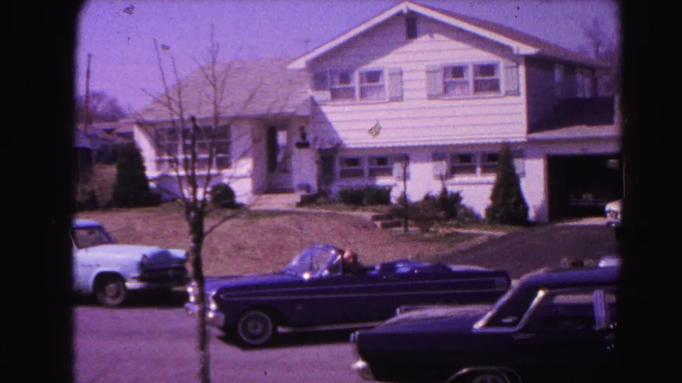 Dark early-1960s convertible pulling further down a suburban street; white two-story house with garage in background; parked cars at curb; Hagerstown, Maryland, 1963 8mm film.