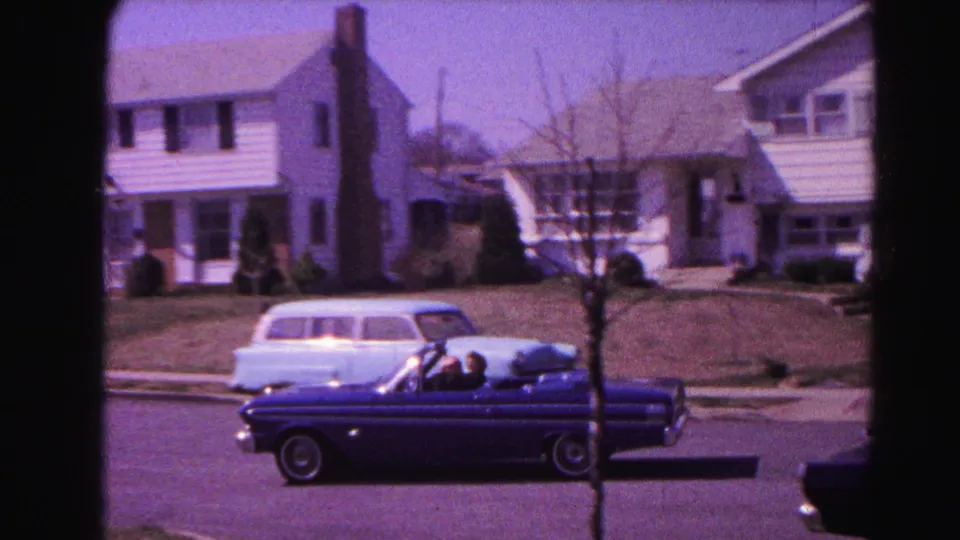 Early-1960s dark blue open-top convertible with two visible occupants passing along a residential street; light station wagon parked at right; neighboring two-story house with chimney; Hagerstown, Maryland, 1963 8mm film.